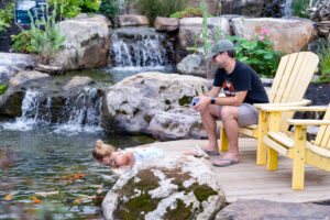 A man sitting in a yellow Adirondack chair while watching his little girl look at the fish in the pond.